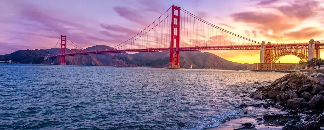 A Ponte Golden Gate ao pôr do sol com um céu multicolorido e a Baía de São Francisco em primeiro plano.